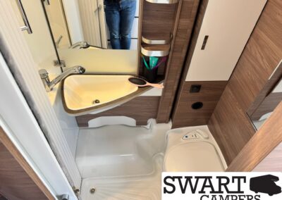 Camper bathroom with white sink, chrome faucet, wood cabinetry, curved shelves, and grooming items in a holder; person’s jeans reflected in the mirror.