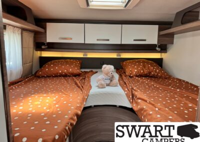 Camper bedroom with two single beds in orange polka-dot sheets, a small teddy bear between them, and white overhead cabinets.
