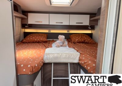 Bunk bed area in a camper with orange polka-dot bed coverings, a teddy bear sitting at the center, white overhead cabinets, and a small ladder reaching to the lower bunk.