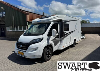 Front-right view of a white KNAUS motorhome with black graphics parked on a brick lot beside an industrial building; blue sky with clouds above. License plate ZJ-632-J visible.