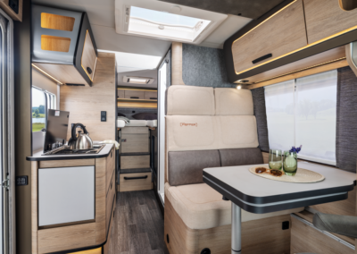 Interior of a modern camper van: kitchenette on the left, a cushioned dinette and table on the right, with a bed area visible toward the back and a skylight above.