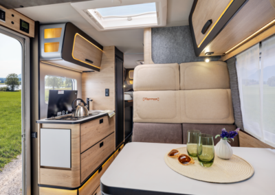 Cozy camper van interior with a compact kitchenette, beige sofa, and a dining table set for two with glasses and a vase.