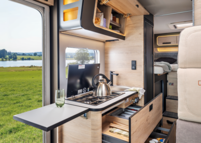 Compact camper van kitchen with a gas stove, kettle, and open wooden shelves; bed area visible in the back and a grassy landscape through the doorway window.