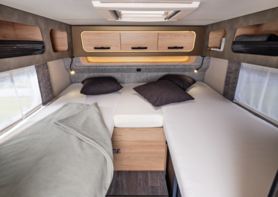 Interior of a compact camper van bedroom with twin bed area, wooden cabinets, and soft pillows beneath a skylight. Note: descriptive but concise alt text conveying layout and amenities.