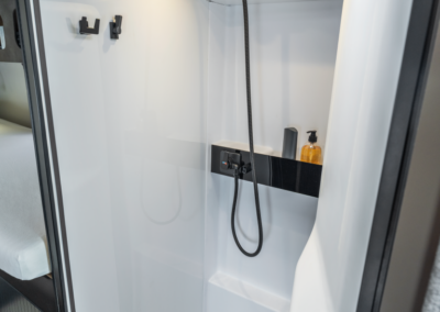 Compact RV shower stall with a handheld shower, black shelf holding a soap dispenser, hooks on the wall, and a skylight above