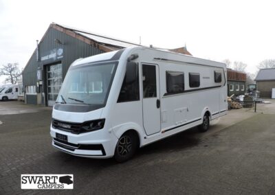 White camper van parked in a lot beside a service workshop, with a Swart Campers logo overlay in the bottom-left corner.