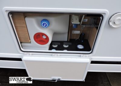 Open exterior cabinet on a white caravan showing a white water tank with a blue fill cap and a red waste cap, wooden interior panels and a small control area inside. Swart Campers logo visible in the lower-left corner.