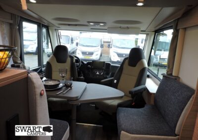Interior of a campervan: front driving seats with a fold-out dining table set for two and a grey sofa along the side, Swart Campers logo visible in the corner