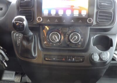 Vehicle dashboard with a large touchscreen infotainment system, climate control dials, gear shifter on the left, and two cup holders below.