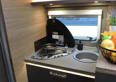 Compact camper kitchen with a two-burner stove, sink, window, and overhead cabinets.