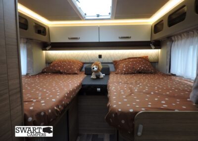 Two twin beds with orange polka-dot bedding in a camper van, a small stuffed lion sits between them under warm lights by a skylight.