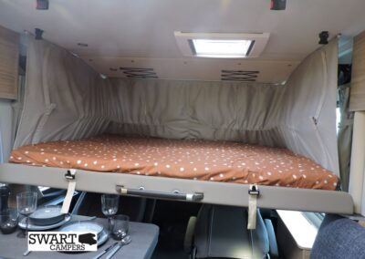 Raised sleeping bunk inside a camper van with brown polka-dot bedding, gray privacy curtains, and a dining table below set with plates and glasses.