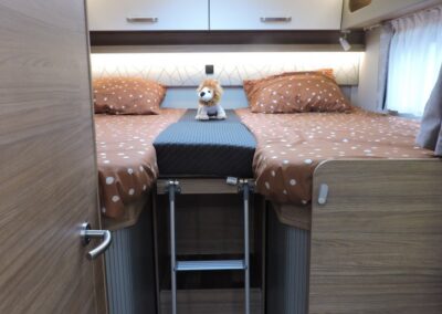 Camper van bunk bed setup with brown polka dot bedding, a small stuffed lion, and a metal ladder to the lower area.