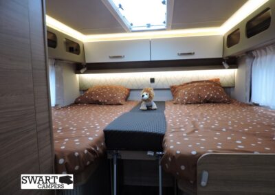 Camper bedroom with two brown polka-dot beds separated by a small plush lion in the center, under warm overhead lighting and a skylight.