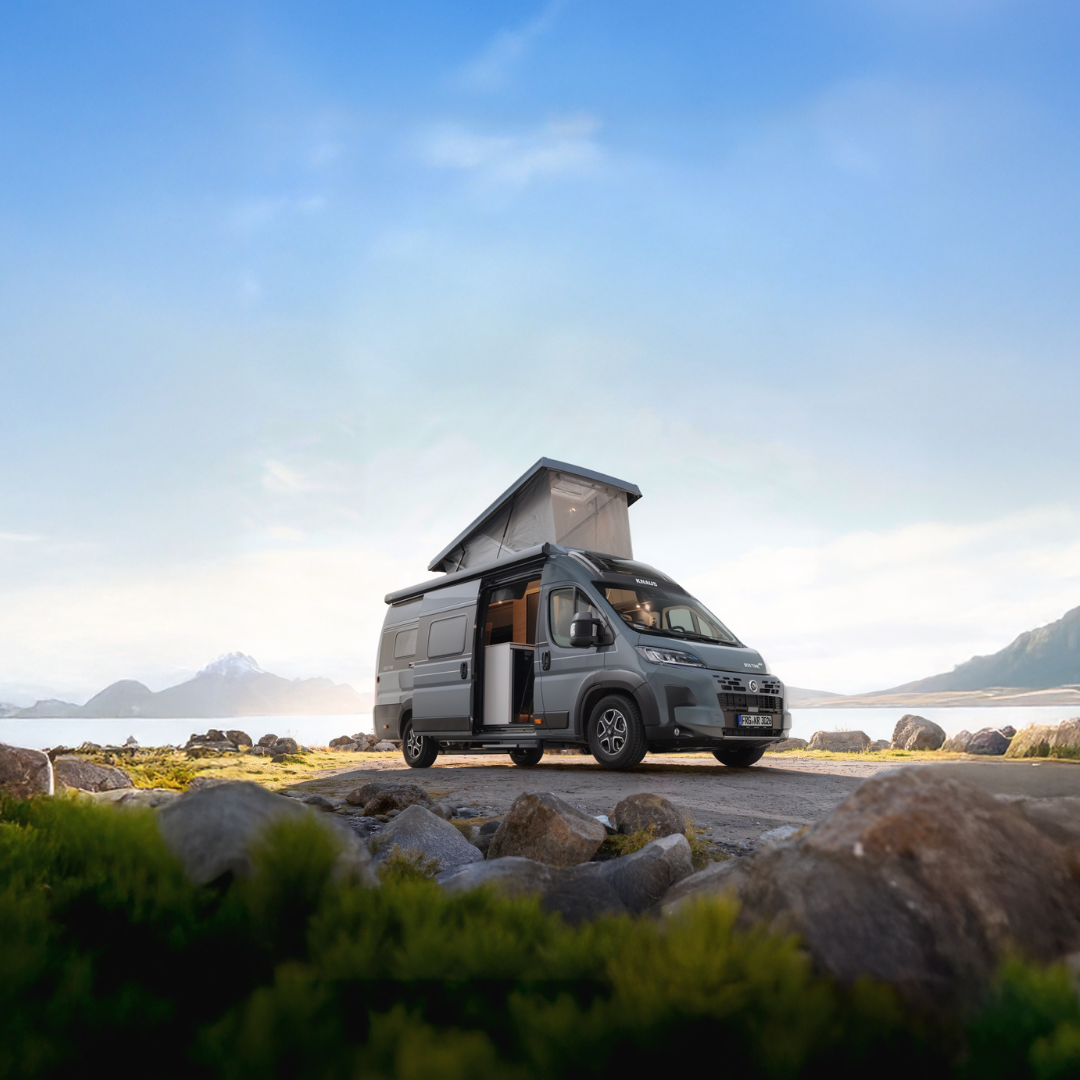KNAUS BOXTIME - Swart Campers Gray camper van with a raised roof tent parked on a rocky lakeshore, mountains in the distance under a blue sky.