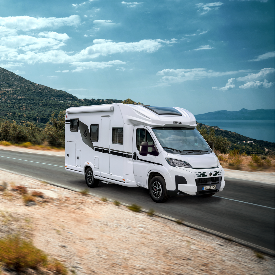 KNAUS LIVE WAVE - Swart Campers White motorhome driving along a coastal highway with hills and sea in the background, under a blue sky with clouds.