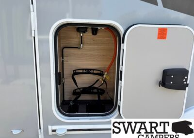 Open RV storage compartment with orange propane hose and gas regulator inside a wood-paneled bay; exterior door open. Swart Campers watermark visible at bottom.
