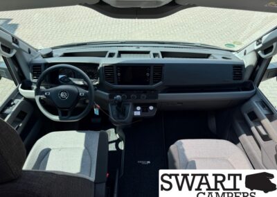 Front cabin of a VW camper van interior, view from the driver's seat with steering wheel, dashboard, and central touchscreen. Swart Campers logo partially overlaid in the corner.