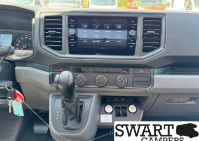 Interior view of a camper van dashboard with a central touchscreen, climate controls, and gear shifter visible.