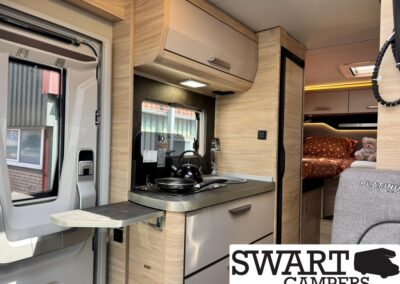Compact camper van interior featuring a small kitchen: fold-out counter, two-burner stove, sink, and light wood cabinetry leading to a sleeping area in back.