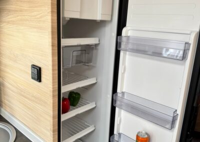 Open camper fridge with shelves, peppers on a lower shelf and a can on the door shelf; compact, built-in unit in a camper kitchen.
