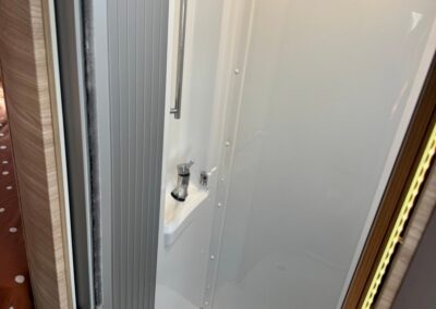 Compact camper shower stall with a chrome vertical shower rail, wall faucet, and sliding gray door, seen from the doorway.