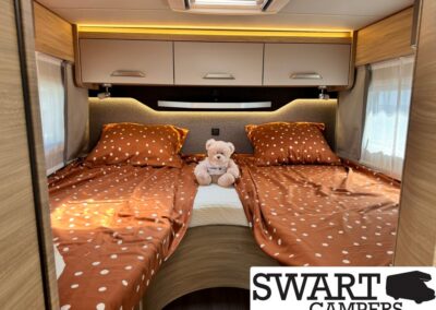 Twin bed setup in a warmly lit camper interior with orange polka-dot bedding and a plush bear between the beds.