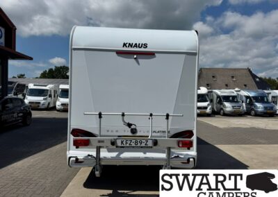 Rear view of a white KNAUS caravan at a dealership, with a bike rack mounted on the back and a Dutch license plate KFZ-89-Z; row of campers in the background and the SWART CAMPERS logo in the bottom-right corner.