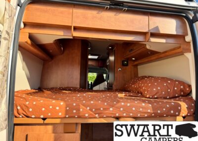 Interior of a camper van showing a raised bed with orange polka-dot bedding, wooden overhead cabinets, and a view toward the cab area at the front. A SWART CAMPERS watermark is visible in the bottom right.