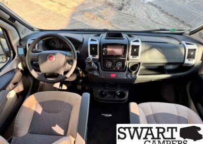 Interior view of a camper van cockpit: steering wheel with Fiat logo, central touchscreen, gear shifter, and two front seats seen from above the dashboard area.