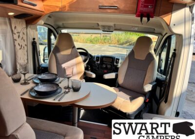 Camper van interior with a round dining table set for two, plates, glasses, and cutlery, front seats visible, SWART CAMPERS logo in corner.