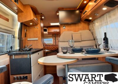 Interior of a motorhome dining area with a round table set for two, beige seating, and a wall-mounted TV.