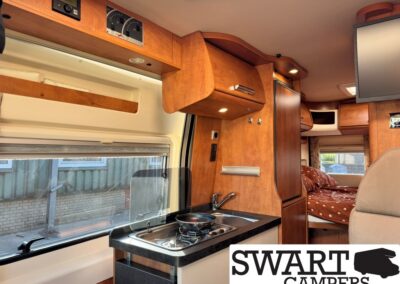 Interior of a wooden campervan: compact kitchen with two-burner stove, sink, and overhead cabinets; sleeping area visible at the back.