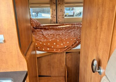 Wood-paneled camper interior featuring a bed with an orange polka-dot duvet, overhead cabinets, and a skylight above.