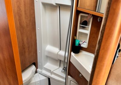 Compact RV bathroom with a white shower stall, handheld shower on a chrome hose, and wooden storage cabinets beside a small sink.