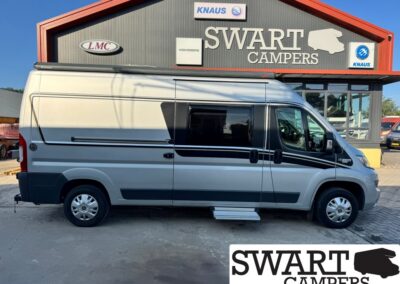 Grey camper van with black stripes parked outside Swart Campers dealership signage visible above the building's entrance in the background.