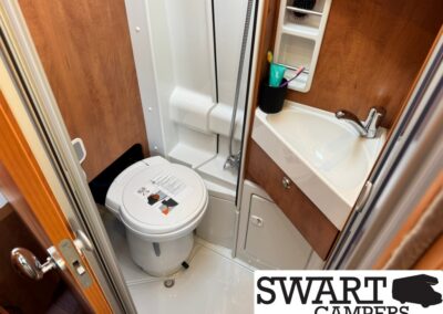 Compact camper bathroom featuring a small toilet, corner shower, and white sink with storage cabinet below.