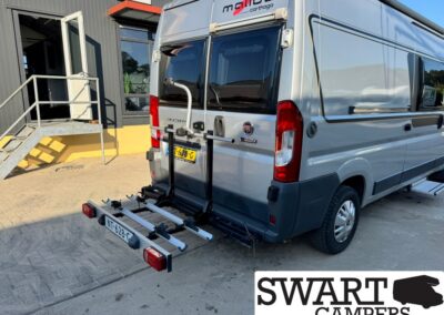 Silver camper van with a rear-mounted bike rack attached, parked outside a building; Swart Campers logo visible in the lower right corner of the image.
