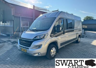 Silver motorhome parked on a brick driveway beside a workshop; Swart Campers logo in the bottom corner.