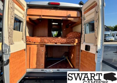 Rear view of a converted camper van interior with a raised bed, orange polka-dot bedding, and wooden cabinetry visible behind open doors for storage and living space.