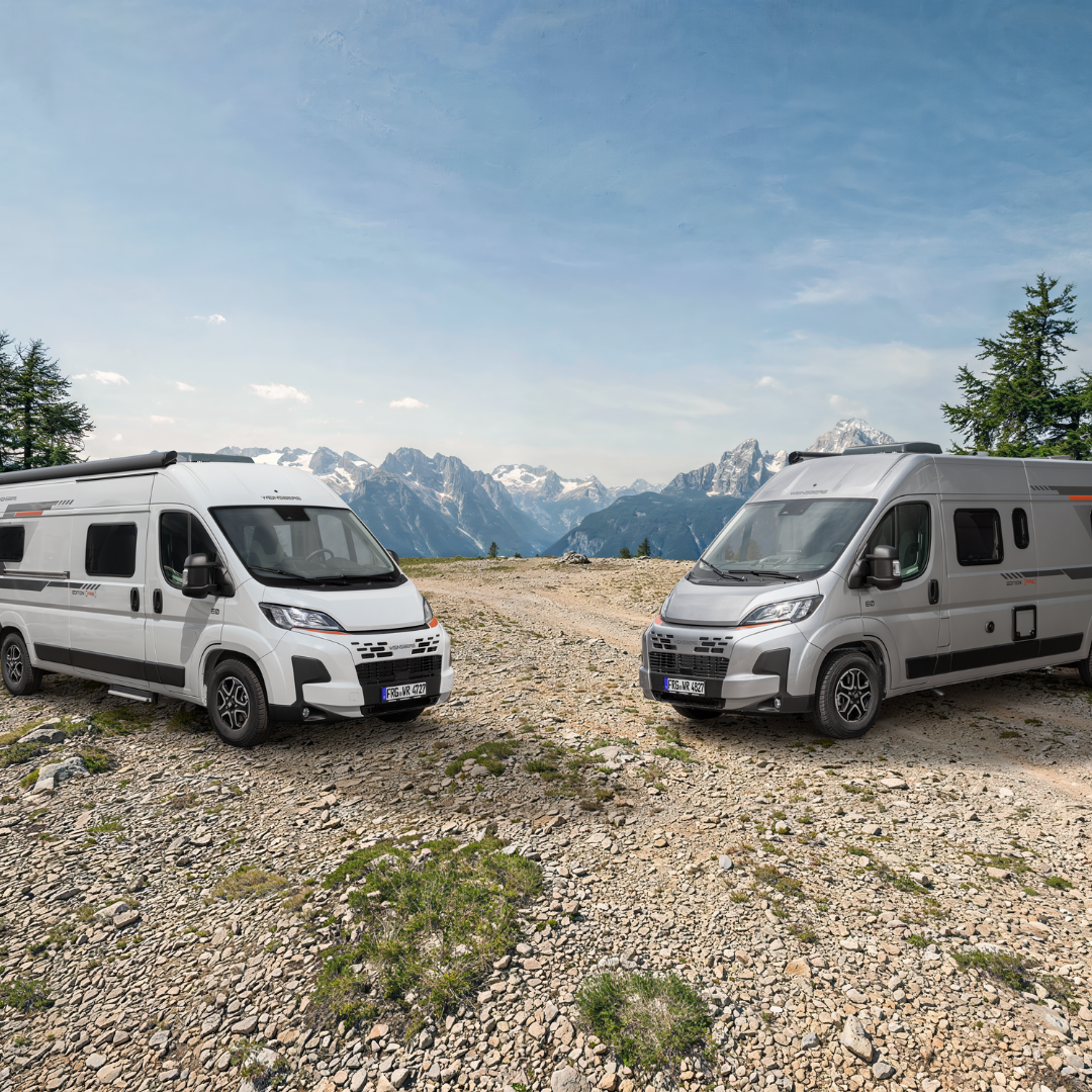 Two white camper vans parked facing each other on a rocky mountain overlook with snow-capped peaks in the distance, under a blue sky.