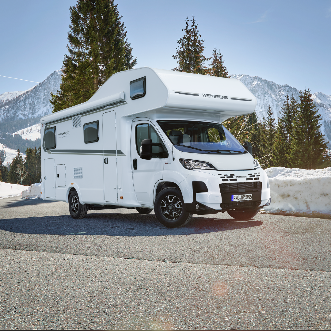 White motorhome parked on a snow-dusted mountain road with evergreen trees and snow-covered peaks in the background.
