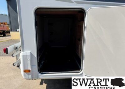 Open storage compartment at the rear of a camper trailer, showing a dark interior and a flat floor with rails along the sides outside a sunny lot.
