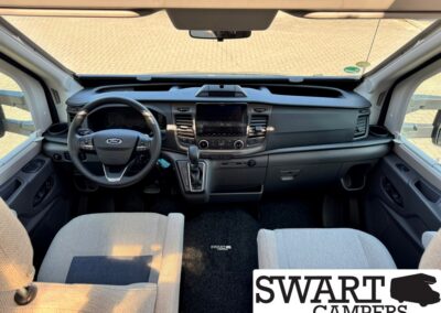 Interior of a camper van cockpit: steering wheel on the left, central touchscreen, climate controls, and gear shifter, with gray front seats and black dashboard.