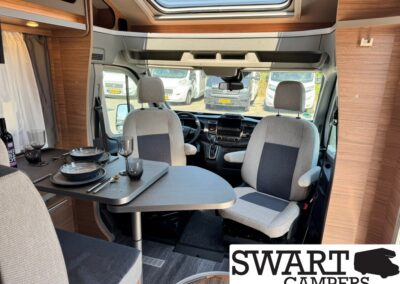 Interior of a camper van with a dining table set for two, gray seats, and the front cabin visible through the windshield window.