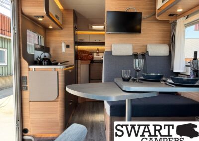 Cozy camper van interior with a dinette, table set for two, kettle on the counter, and a wall-mounted TV; wood paneling and a bed in the back. SWART CAMPERS logo visible in the corner.
