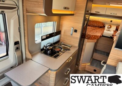 Cozy camper kitchen with a two-burner stove and kettle, fold-out counter, and wooden cabinets, leading to a bed area.