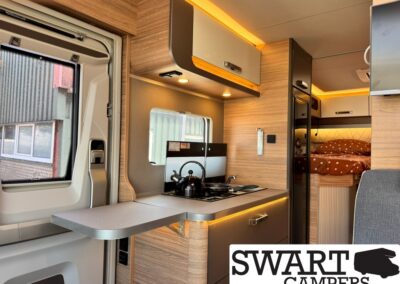 Compact camper van kitchenette with a two-burner stove and kettle on the counter, wood cabinets, and a bed visible in the rear area.