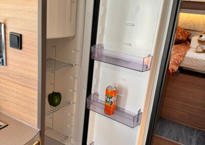 Open compact refrigerator in a camper, with an orange Fanta can on the door shelf and a green bell pepper on the middle shelf; bed and warm wood interior visible in the background.