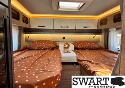 Cozy camper bedroom with two single beds in brown polka-dotted sheets and a small stuffed lion between them, under warm overhead lighting.